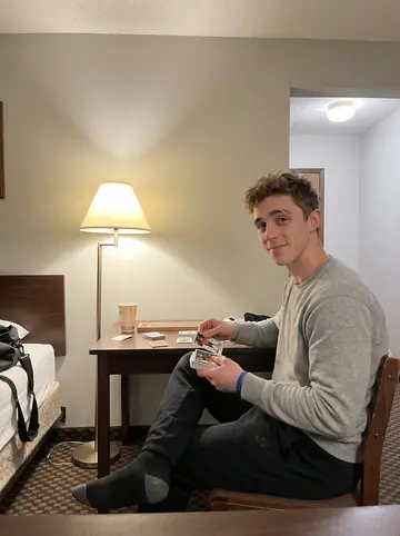 Ronald, AI character: budget motel room on a road trip, small desk covered with hockey cards, a cribbage board, and a half-empty…