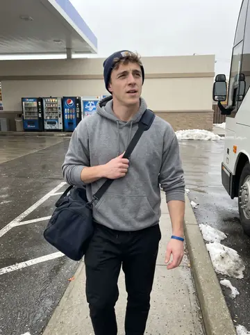Ronald, AI character: gas station parking lot before the team bus leaves, wet pavement and a row of vending machines behind him