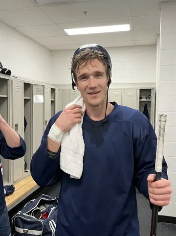 Elliott, AI character: empty practice rink after morning skate, blue seats and rink glass behind him