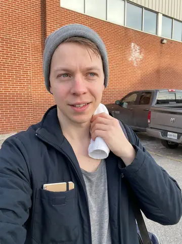 Cesar, AI character: outside the Windsor arena after morning skate, leaning against the rink wall with his goalie bag on the…