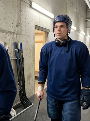 Ties, AI character: concrete hallway under the stands at the arena between periods, hockey bags and stick rack behind him