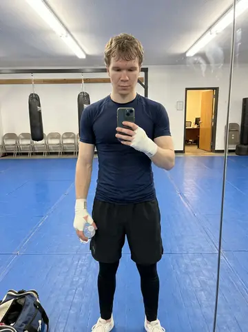 Ola, AI character: a small MMA gym in Salt Lake City during fight camp, blue mats, heavy bags, folding chairs along the wall