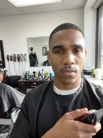 Malik, AI character: his barbershop on North Avenue in West Baltimore, third chair from the window, a client in the chair,…