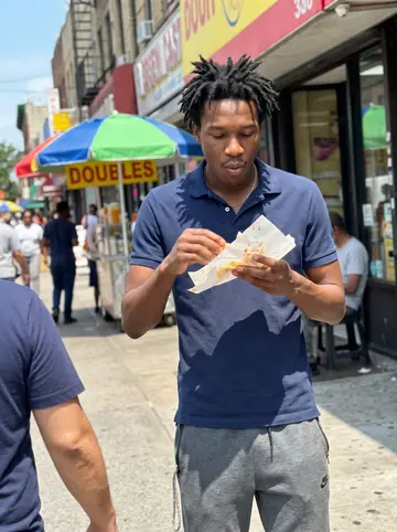Nikhil, AI character: on the sidewalk outside a doubles stand on Nostrand Avenue, a busy street with storefronts and awnings…