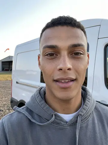 Zion, AI character: the gravel lot beside a drop zone in south Georgia, leaning on the sliding door of his white cargo van, a…