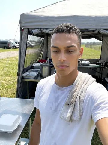 Zion, AI character: the open-sided camp kitchen tent at the drop zone, a steel prep table with foil trays, a big pot of red…