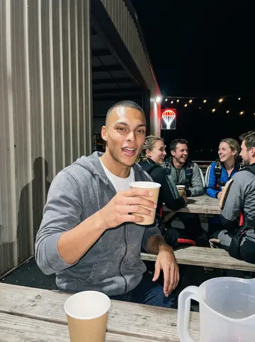 Zion, AI character: wooden picnic tables outside the drop-zone hangar after the last load, string lights overhead, other…