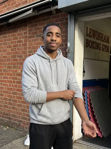 Nigel, AI character: outside the Lewisham boxing gym after the Saturday kids' class, red brick wall and gym doorway behind him