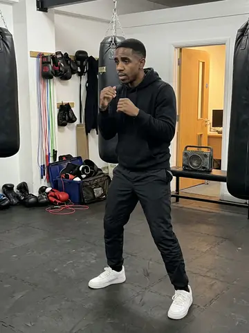 Nigel, AI character: inside the Lewisham boxing gym beside the heavy bags, kids' gloves and skipping ropes piled near the wall