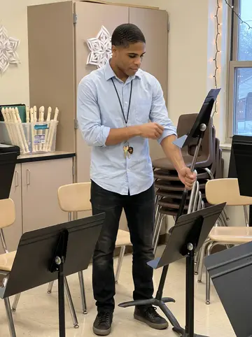 Kwadwo, AI character: empty elementary school music room after dismissal, little chairs stacked, recorders in a plastic bin,…