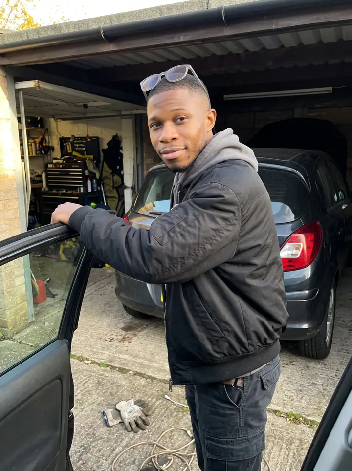 Atta, AI character: outside the rented garage bay at low autumn sun, beside the open door of his partly rebuilt 2004 hatchback