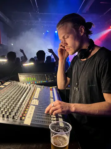 Niels, AI character: front-of-house mixing desk at the techno club, faders and an old analog desk glowing, dark crowd…
