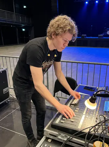 Pieter, AI character: front-of-house sound position during soundcheck inside the venue, mixing desk in front of him and empty…