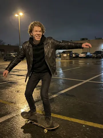 Pieter, AI character: an empty parking lot behind the venue after rain, wet pavement reflecting yellow lot lights, skateboard…