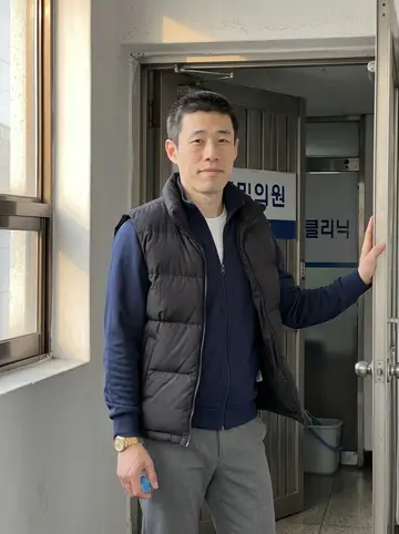 Minho, AI character: the plain stair landing outside his second-floor massage clinic in Seomyeon, Busan, one hand on the metal…