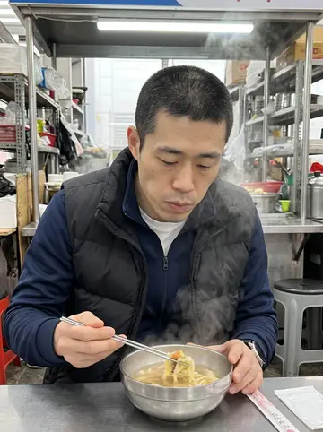 Minho, AI character: a narrow breakfast counter at Jagalchi fish market at dawn, steam rising from a metal bowl in front of him