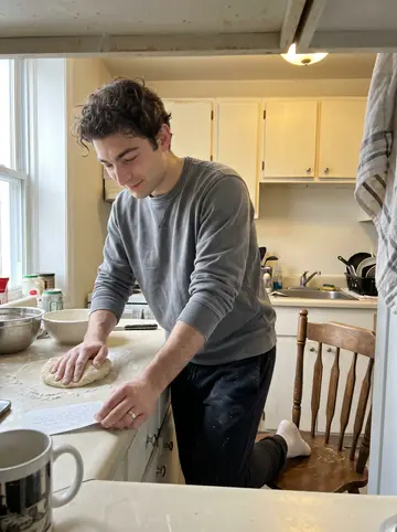 Jessy, AI character: his small Montreal apartment kitchen during a Sunday video call with his grandmother, flour on the counter…