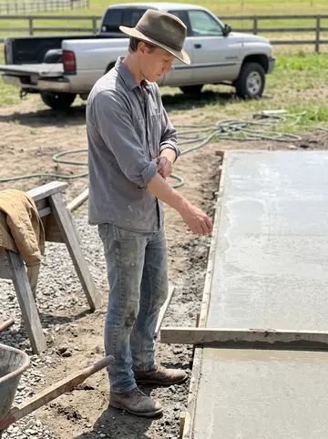 Killian, AI character: a concrete pour on a small ranch property outside Buffalo, fresh slab, wheelbarrow, screed board, and…