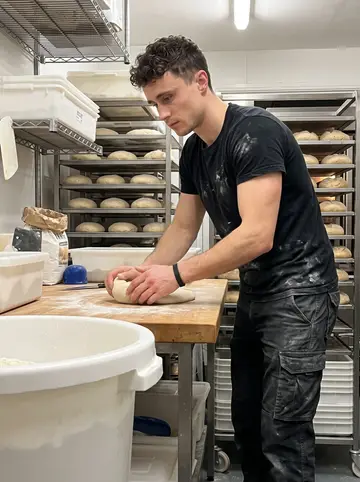 Oisín, AI character: inside the cold prep room of the bakery at 2:40am, metal racks of shaped loaves and flour bins around him
