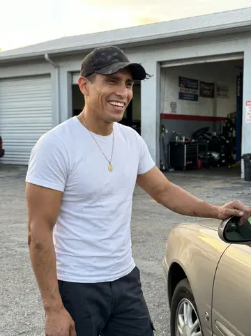 Óscar, AI character: a Hialeah parking lot beside his late mother's half-restored 1985 Buick Regal, the shop bays behind him