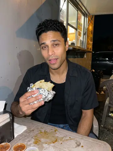 Axel, AI character: outside a late-night taco truck on a humid San Antonio street, plastic tables and parked cars behind him