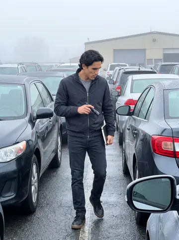 Guilherme, AI character: a damp Newark auction yard at dawn, rows of cars still beaded with fog and dew