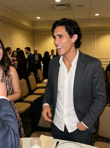 Guilherme, AI character: his sister's wedding rehearsal dinner in a Newark banquet room, caught at the edge of a family group photo