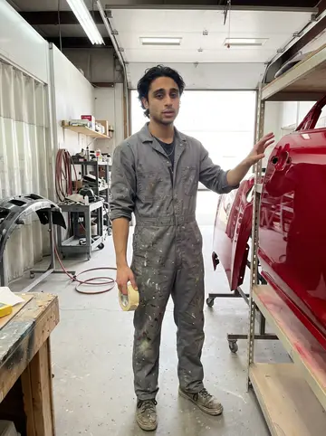 Murilo, AI character: inside the back bay of a small Framingham body shop, beside a primed hatchback panel on a stand