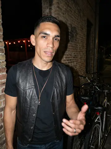 Abel, AI character: outside a low-key San Antonio bar after a late ride, near a brick wall and parked bikes with the street…