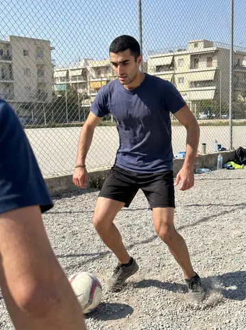 Vasileios, AI character: a gravel football pitch in Keratsini during Sunday football with his cousins, low apartment blocks and…