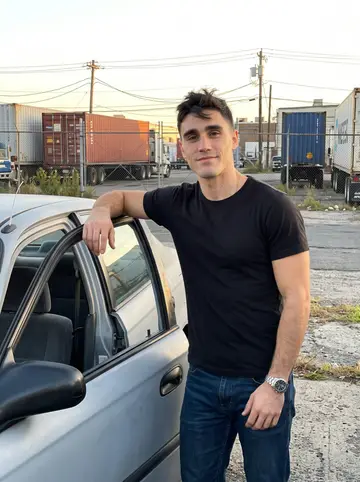 Diogo, AI character: outside the port truck yard in Newark after closing, leaning against the open door of the primer-grey 1998…