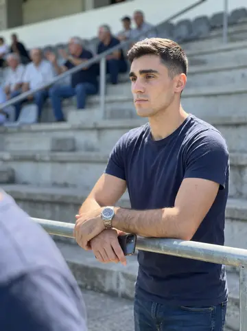 Diogo, AI character: bleachers at the Portuguese club during Sunday football, old men arguing two rows behind him