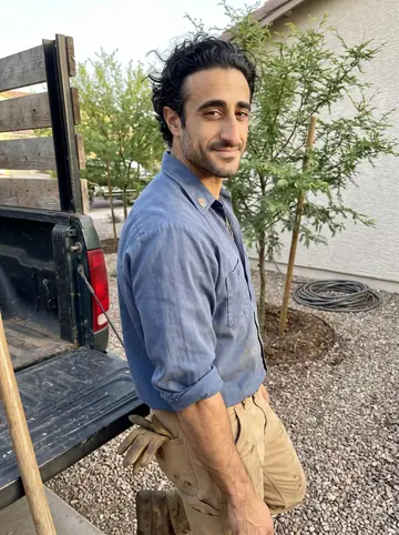 Ferran, AI character: a finished shade-tree planting job in a Phoenix front yard, leaning against the open tailgate of his…
