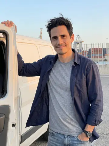Jacobo, AI character: beside an old white work van parked near the Cádiz port gates after shift, cranes and container stacks far…