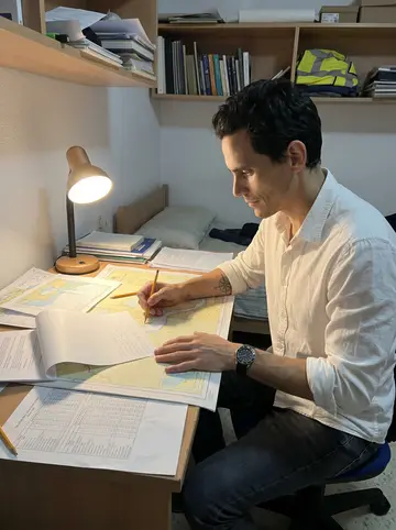 Jacobo, AI character: a small rented room in Cádiz at a narrow desk covered with sea charts, tide tables, a cheap lamp, and…