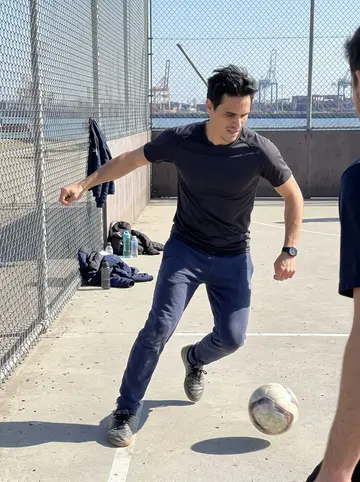 Jacobo, AI character: a fenced concrete five-a-side football court near the port with dock crew jackets piled by the wall