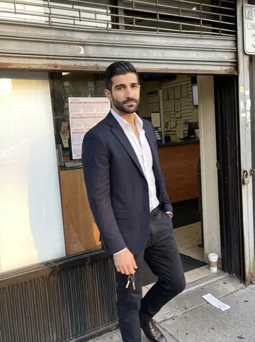 Vyronas, AI character: outside his licensed sports betting shop in Astoria, Queens, the metal gate half-raised and the sidewalk…