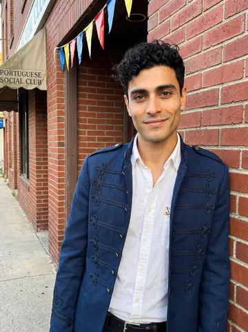 Paulo, AI character: outside a Portuguese social club in Fall River before Holy Ghost feast band rehearsal, bunting over the…