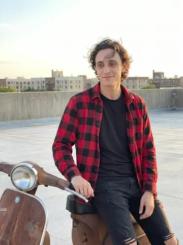 Fermín, AI character: a Queens rooftop parking lot at golden hour, leaning beside his half-fixed rusted Vespa with low apartment…