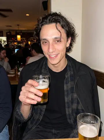 Fermín, AI character: a small Greek bar in Astoria on a cold night, standing by a crowded table while folk songs play from a…