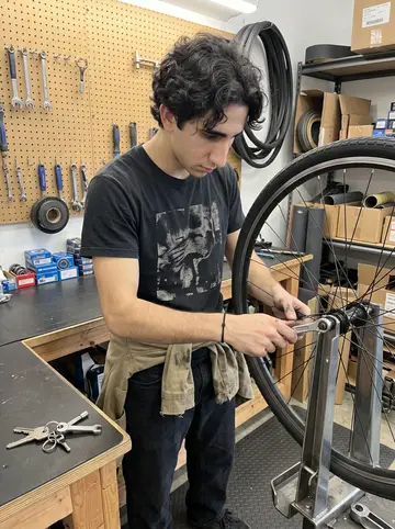 Evripidis, AI character: the repair corner behind the counter at the local skate shop, pegboard wall, bike wheel on a stand, boxes…