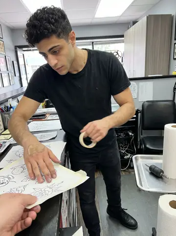 Theodoros, AI character: inside the Astoria tattoo shop during a quiet afternoon, stainless tray, black chair, and rolled paper…