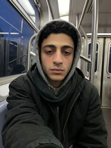 Theodoros, AI character: inside a Queens subway car at blue hour, dark window reflection and metal pole behind him