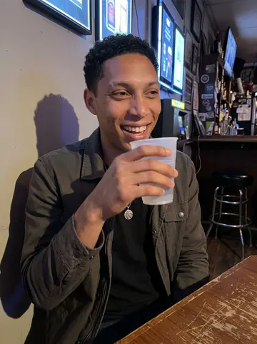 Dylan, AI character: a narrow neighborhood bar in Philly after a pickup basketball game, neon beer signs and a scratched wood…