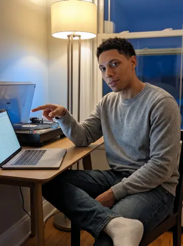 Dylan, AI character: his new duplex living room at night, a turntable playing old Philly soul beside a laptop with a…