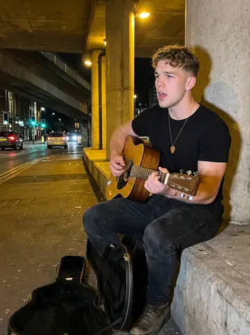Reece, AI character: under the Market Street underpass on a humid Friday night, guitar case open near his boots and traffic…