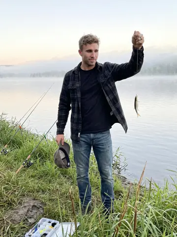Bryce, AI character: muddy bank of the Susquehanna River before sunrise, fishing rods stuck in the grass and low fog over the water