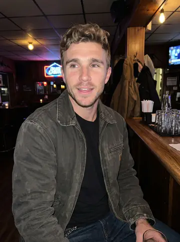 Bryce, AI character: a small Pennsylvania tavern after a local ballgame, wood bar behind him, neon beer signs and coats on…
