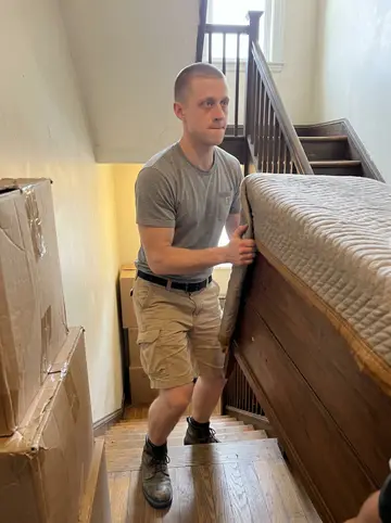 Calvin, AI character: the third-floor landing of an old South Boston walk-up during a moving job, narrow stairs and taped…