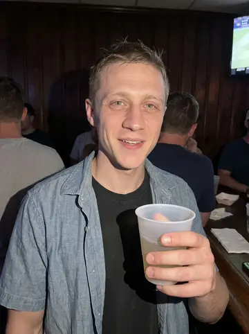Calvin, AI character: a crowded neighborhood bar after a Celtics win, dark wood wall and a TV glow behind him